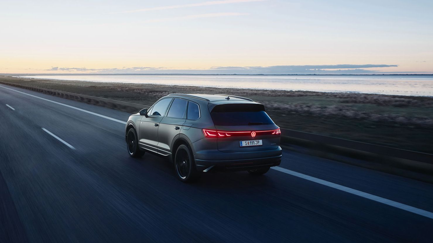 Ein dunkler Touareg R fährt auf einer leeren Straße entlang der Küste bei Sonnenuntergang. Die Straße erstreckt sich in die Ferne, während der Himmel im Hintergrund mit warmen Farben erleuchtet ist. Links und rechts der Straße befinden sich Wasserflächen und Grünflächen.