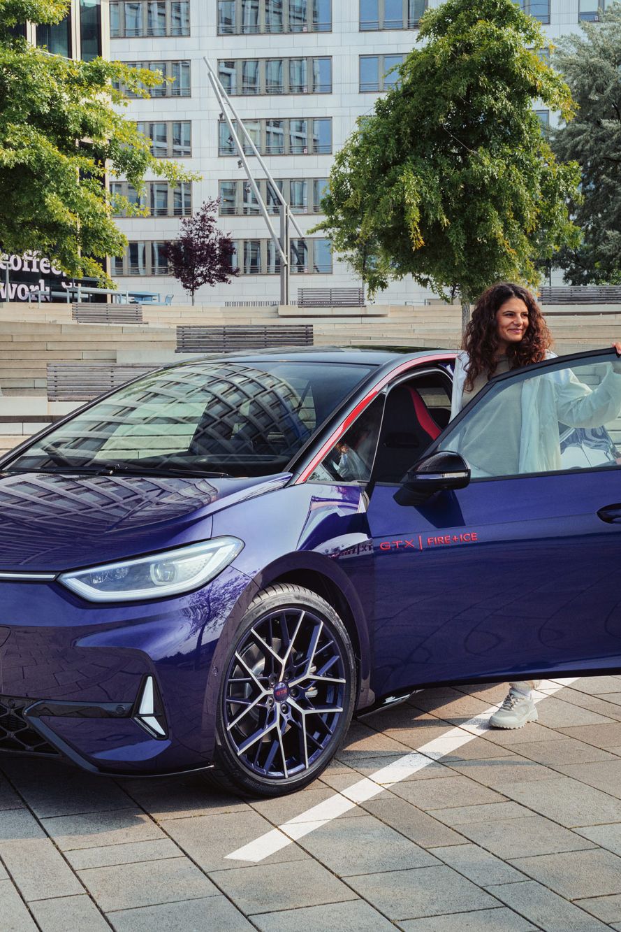 Eine Person steigt aus einem dunkelblauen VW Elektroauto auf einem modernen Parkplatz aus. Im Hintergrund sind Treppen und grüne Bäume zu sehen. Im Vordergrund des Autos sind Markierungen auf dem Asphalt sichtbar.