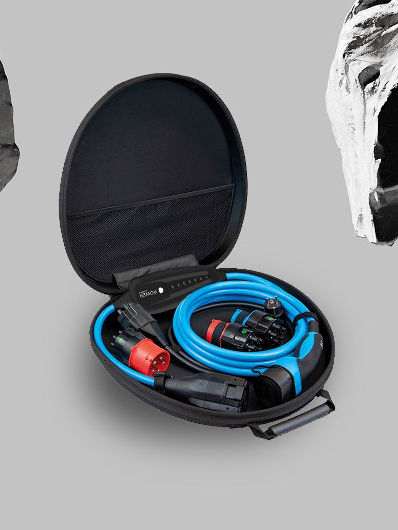 Ein offener, ovaler Koffer enthält blaue und schwarze Kabel sowie Anschlüsse. Der Hintergrund ist grau und mit schwarz-weißen Gesteinsformen verziert. Das Bild zeigt Ausrüstung für den Anschluß von Elektrofahrzeugen.