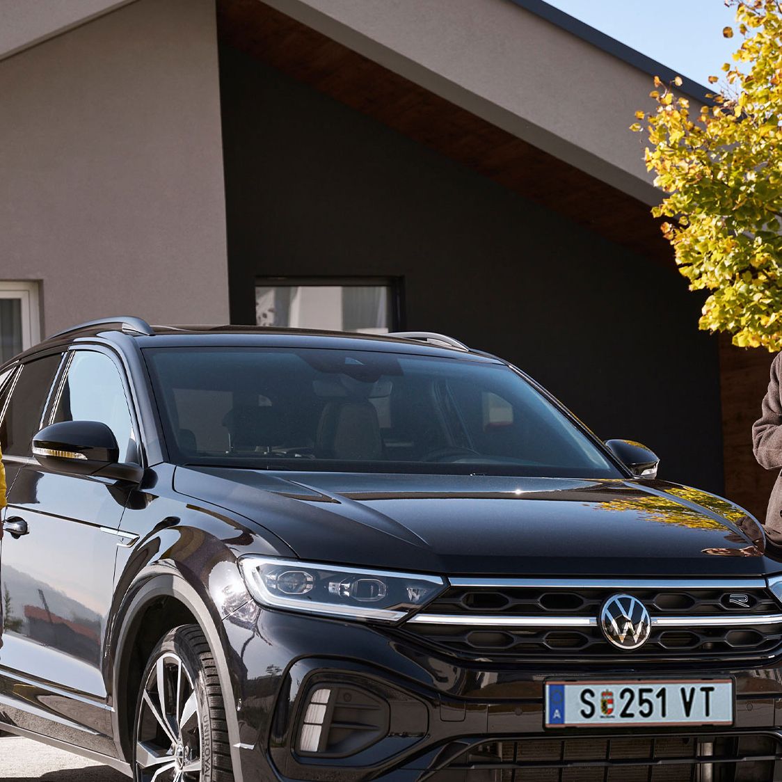 Mann und Frau steht mit Smartphone neben einem geparkten T-Roc vor einem Haus.