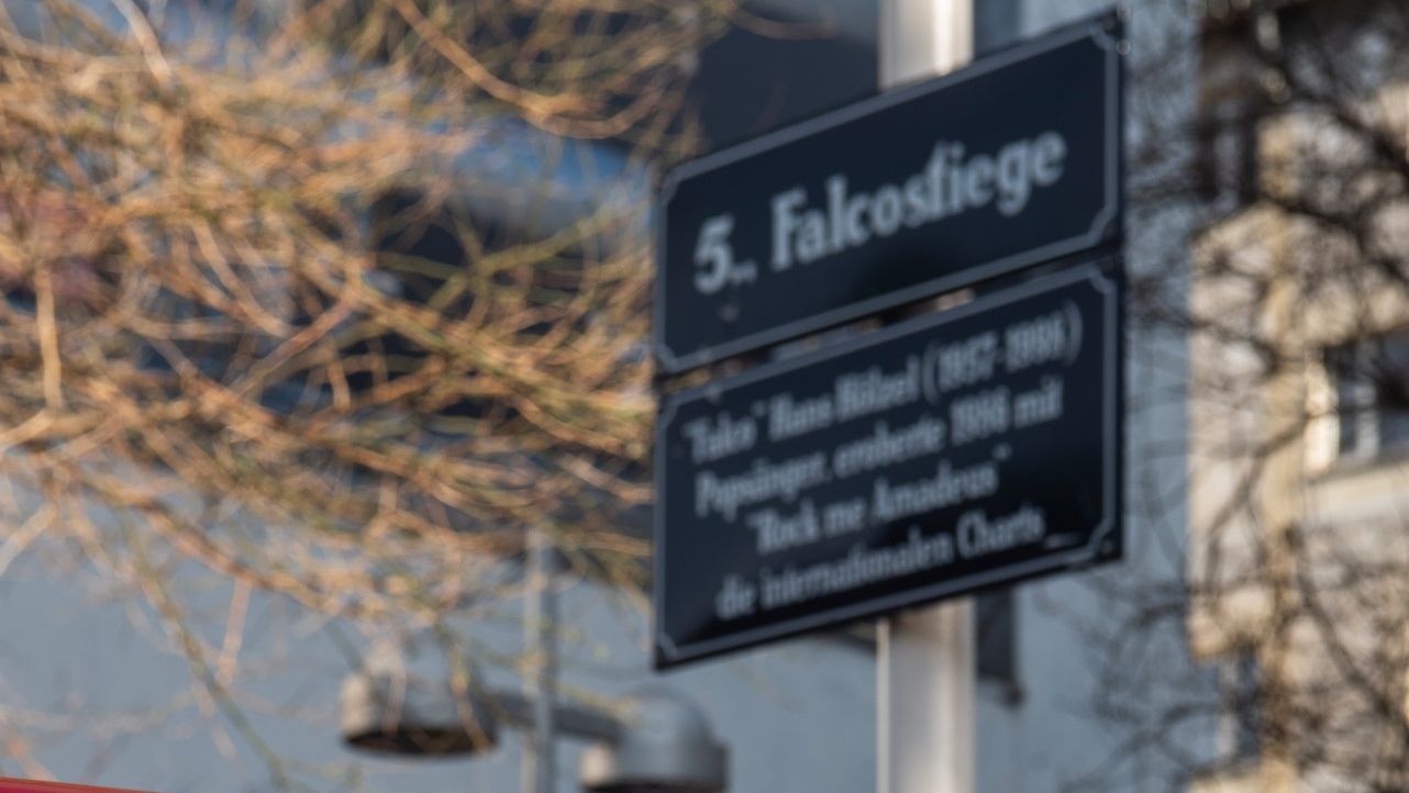 Nahaufnahme der Rückseite eines roten Autos mit der Aufschrift „Falco Edition“ und der stilisierten Unterschrift „Falco“ auf dem Lack. Im unscharfen Hintergrund steht ein Straßenschild mit der Bezeichnung „5. Falcosiege“ und einem Hinweistext, teilweise von Ästen verdeckt.