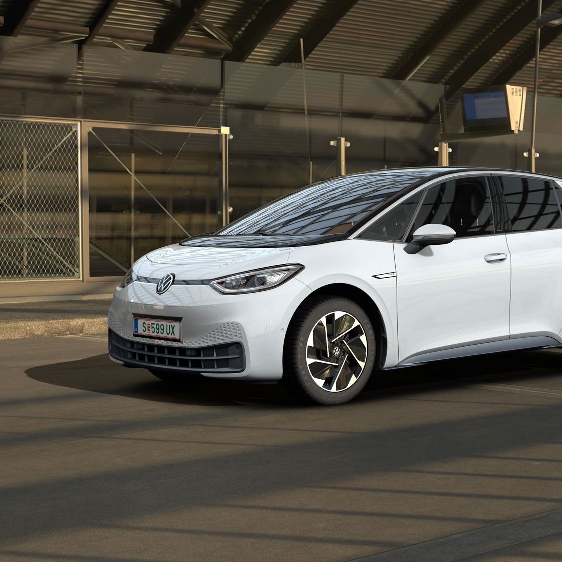 Ein weißer Volkswagen ID.3 steht in einer modernen, teilweise verglasten Tiefgarage. Das Auto ist von der Seite und leicht von vorne zu sehen. Im Hintergrund sind Maschendrahtzäune und Betonwände zu erkennen.