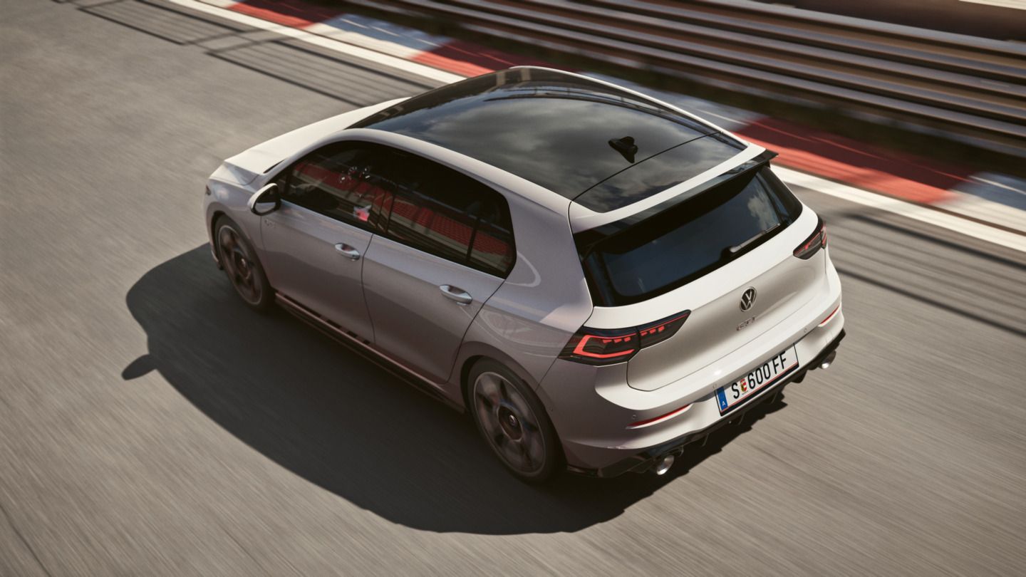Ein silberfarbener Volkswagen Golf GTI fährt schnell auf einer Rennstrecke. Die Perspektive ist von oben und hinten, die Felgen sind schwarz und das Fahrzeug hat ein Dachfenster. Im Hintergrund sind rote und weiße Kurvenmarkierungen zu sehen.