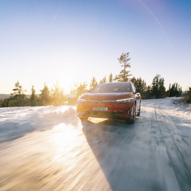 Ein roter ID.7 fährt auf einer eisigen Straße bei Sonnenaufgang. Im Hintergrund sind verschneite Wälder und Berge zu sehen. Die Sonne scheint hell und wirft lange Schatten.