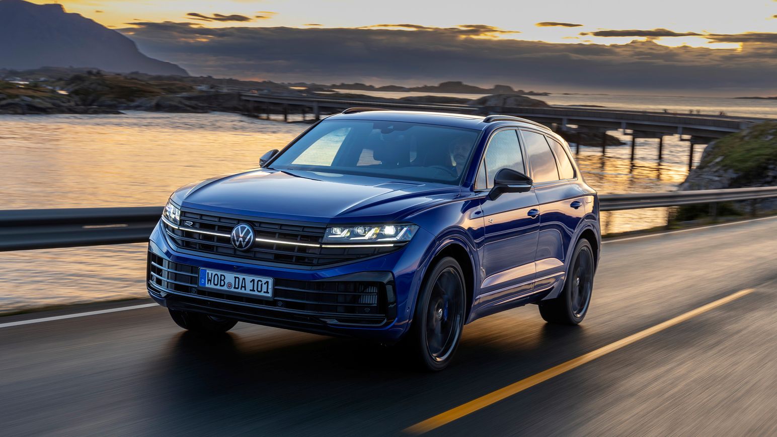 Ein blauer VW Touareg fährt bei Sonnenuntergang neben dem Meer.