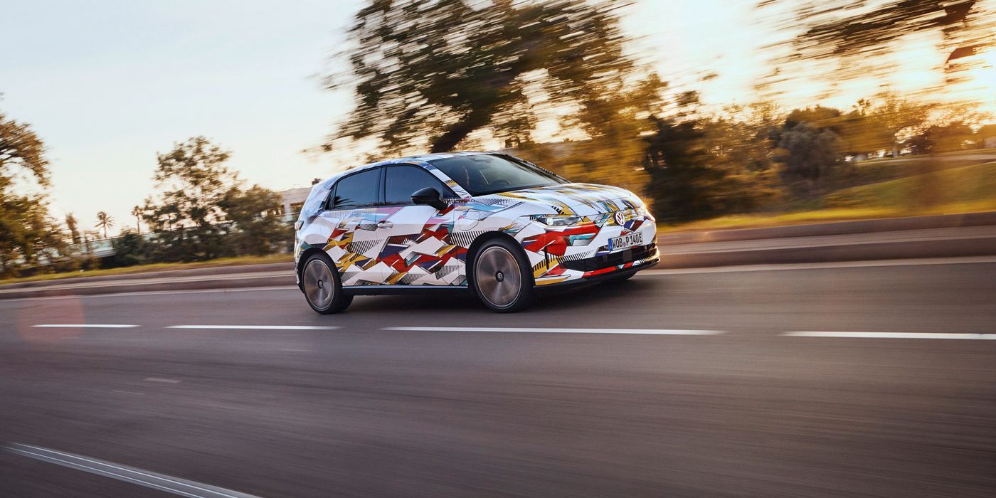 Ein ID. Polo fährt schnell auf einer Straße, umgeben von verschwommenen Bäumen und Landschaft bei Sonnenuntergang. Das Fahrzeug trägt eine auffällige, bunte Tarnlackierung. Die Straße ist leer und scheint sich durch eine ländliche Gegend zu schlängeln.