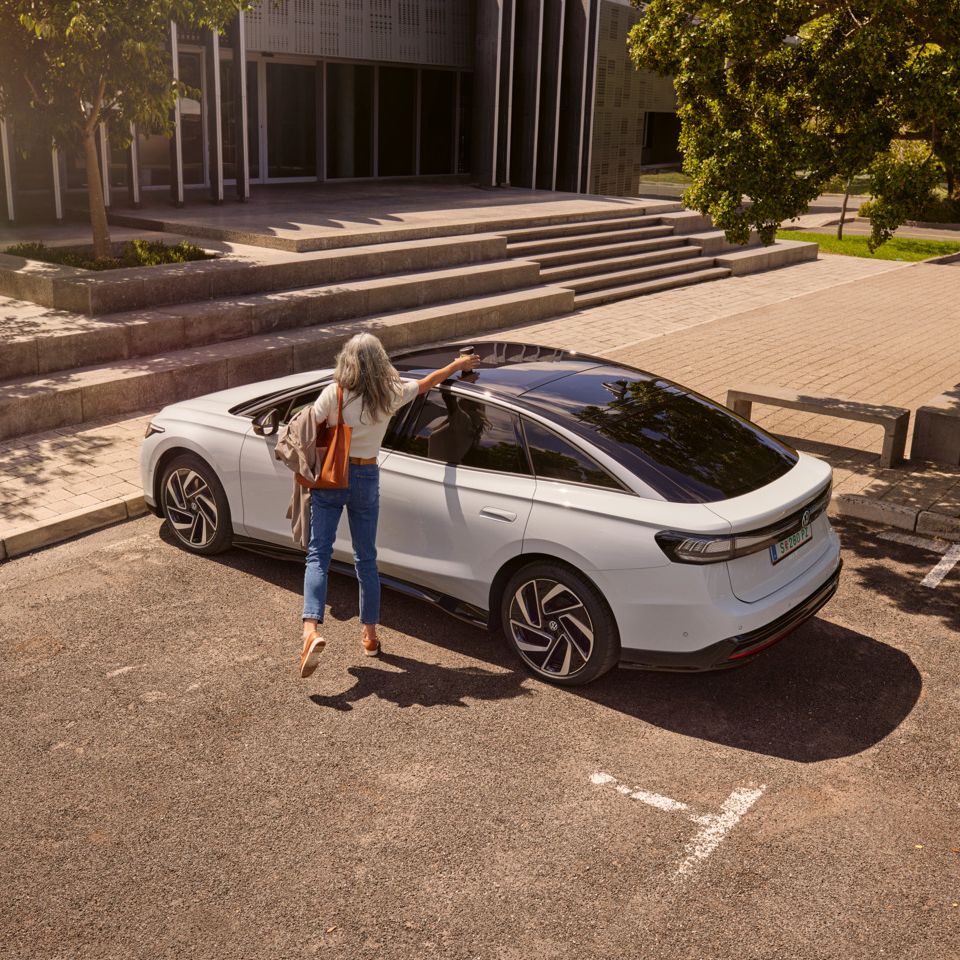 Eine Frau nimmt einen Becher vom Dach eines VW ID.7, der vor einem Bürogebäude parkt.