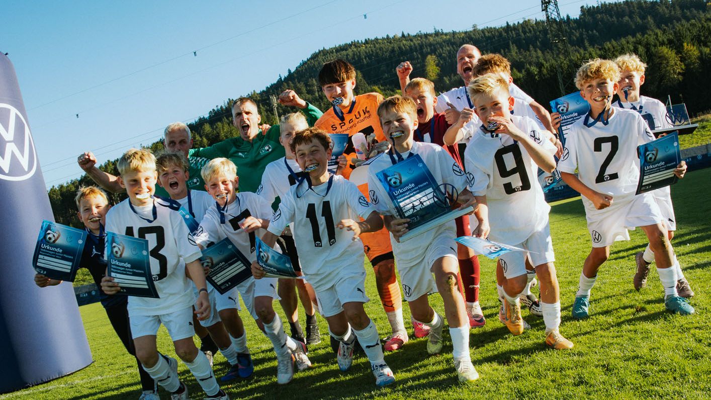 Eine Gruppe jubelnder Kinder in weißen Fußballtrikots läuft auf einem Rasenplatz direkt auf die Kamera zu. Viele von ihnen halten Urkunden oder Pokale in den Händen, einige springen vor Freude in die Luft. Im Hintergrund sind Trainer, ein großes Werbeschild und eine bewaldete Hügellandschaft unter blauem Himmel zu sehen.