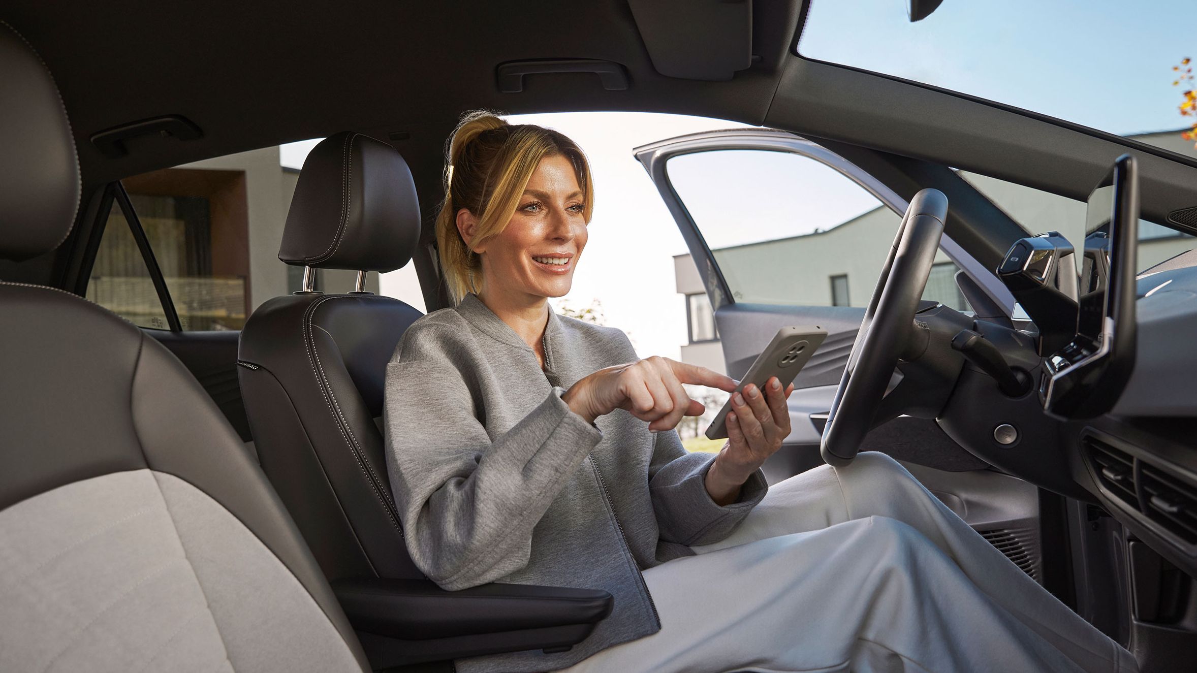 Eine Frau sitzt in einem Volkswagen auf dem Fahrersitz und bedient ein Smartphone. Der Innenraum des Autos wirkt hochwertig. 