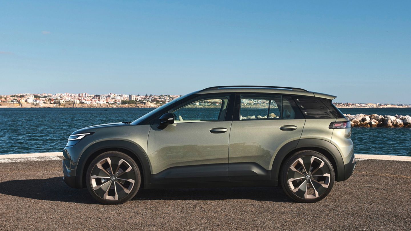 Ein grüner VW ID. Cross steht auf einer gepflasterten Fläche vor einem Meer, das von einem klaren blauen Himmel überdacht ist. Im Hintergrund sind Gebäudestrukturen sichtbar.