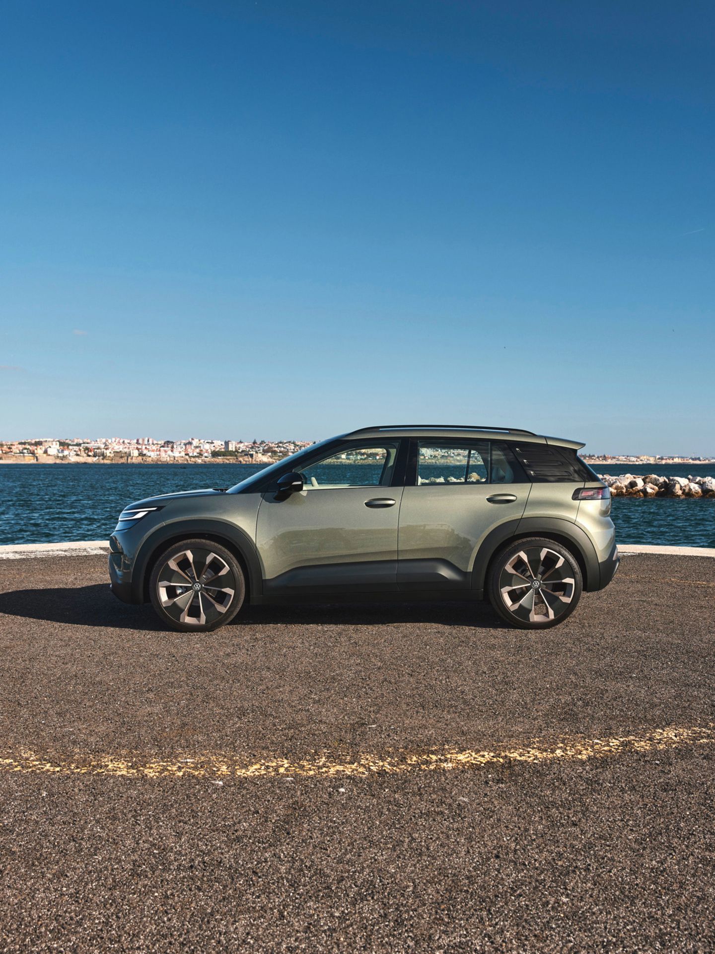 Ein grüner VW ID. Cross steht auf einer gepflasterten Fläche vor einem Meer, das von einem klaren blauen Himmel überdacht ist. Im Hintergrund sind Gebäudestrukturen sichtbar.