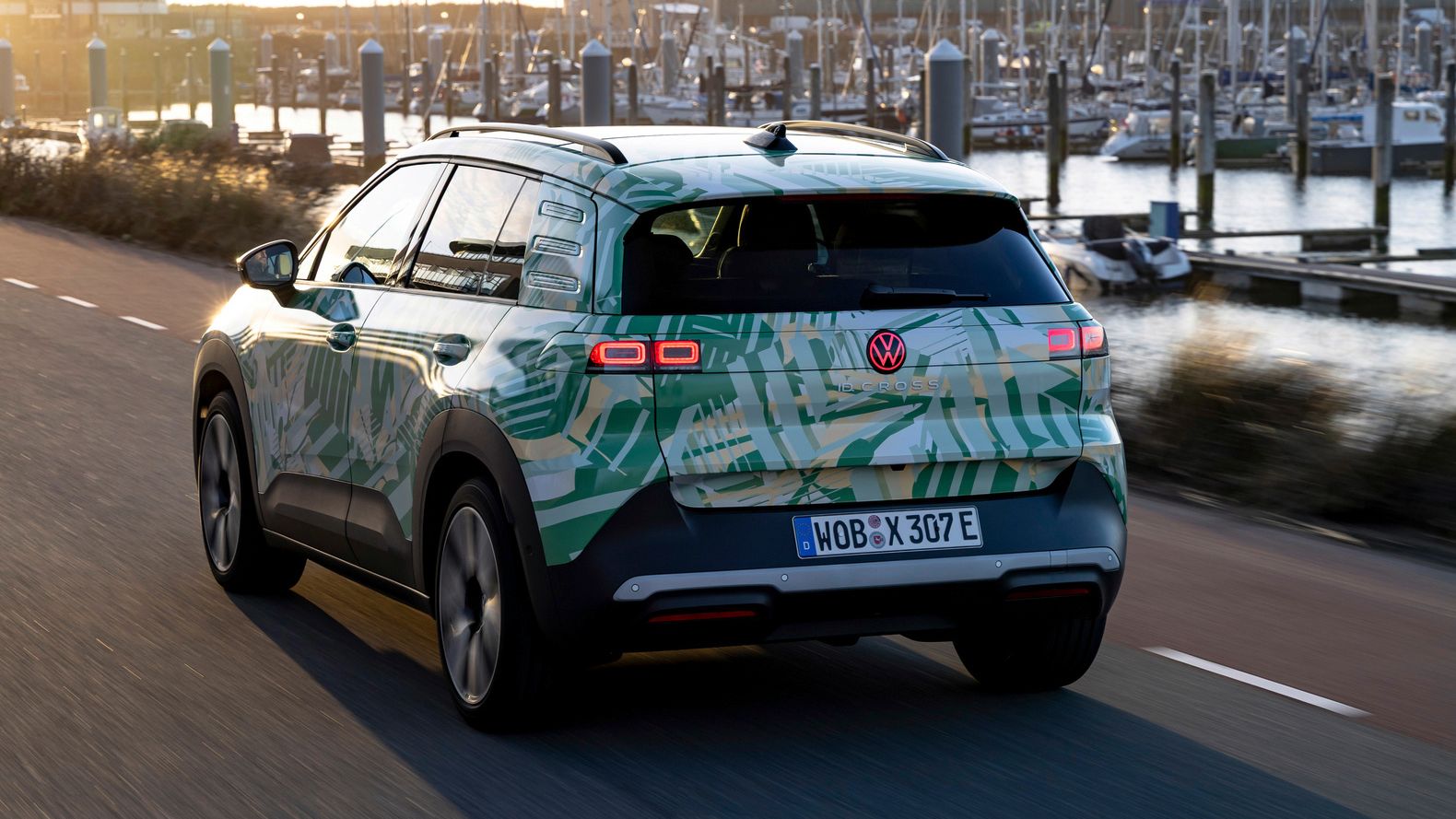 Ein Volkswagen-Elektroauto fährt auf einer Straße entlang eines Hafens bei Sonnenuntergang. Im Hintergrund sind viele Segelboote und Yachten zu sehen. Die Straße und die Landschaft wirken ruhig und friedlich.