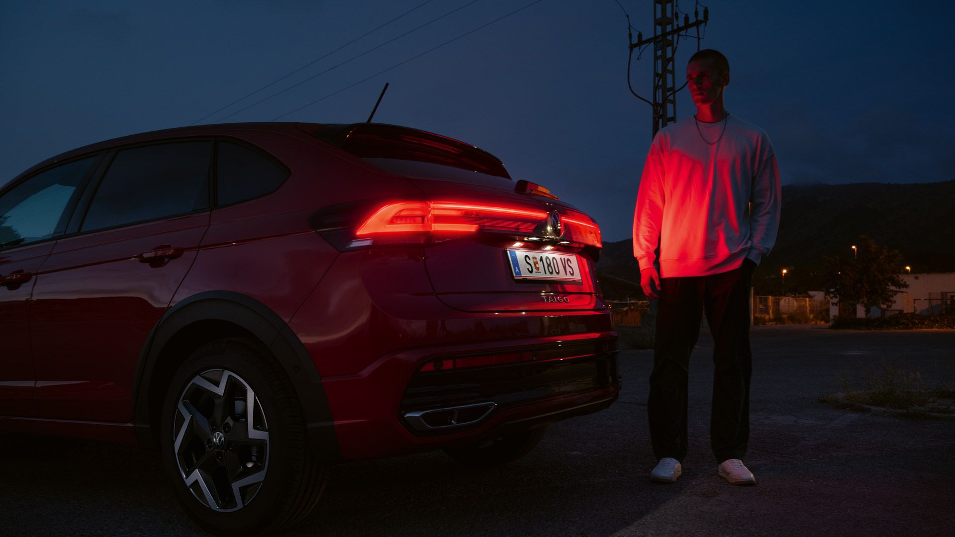 Ein Mann steht am Heck eines VW Taigo R-Line im Dunkeln und wird von den Rückleuchten rot angeleuchtet.