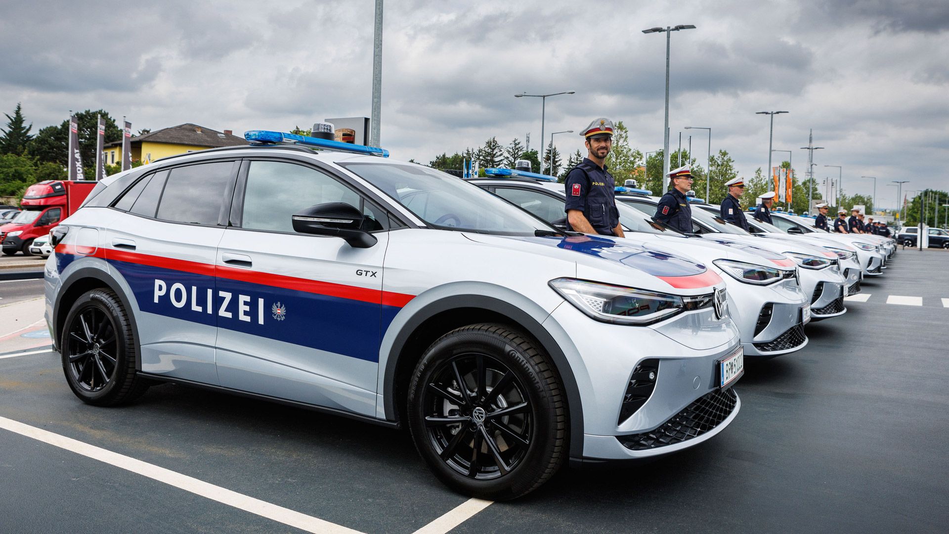 Eine Reihe von Polizeifahrzeugen ist geparkt, es sind silberne ID.4 GTX mit blauen und roten Streifen. Im Vordergrund steht ein Polizist neben einem Fahrzeug. Der Himmel ist bewölkt.