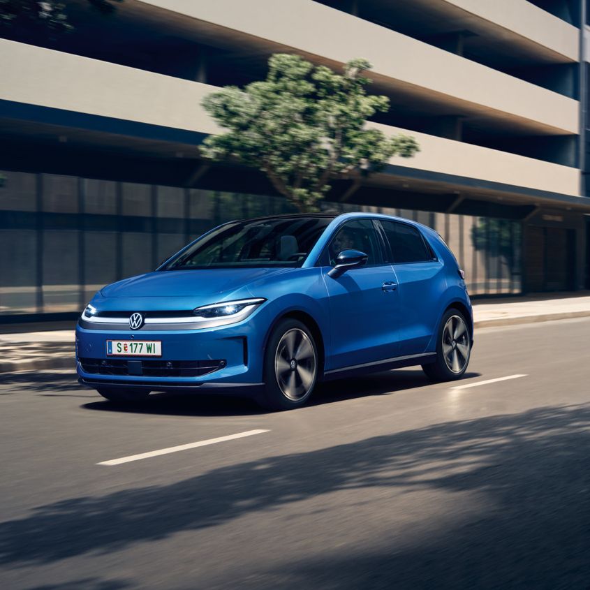 Ein blauer VW ID. Polo Style fährt durch eine Stadt mit grünen Bäumen am Straßenrand.