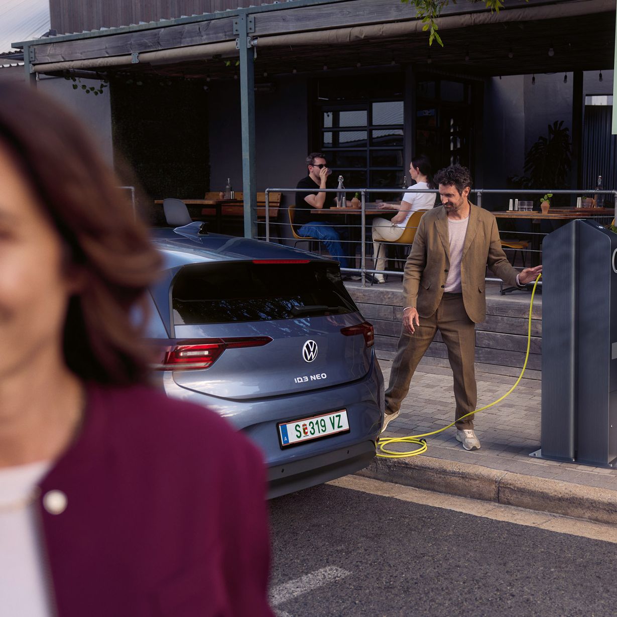 Eine Frau geht auf einer Straße. Hinter ihr läd ein Mann einen VW ID.3 Neo an einer Ladesäule vor einem Café