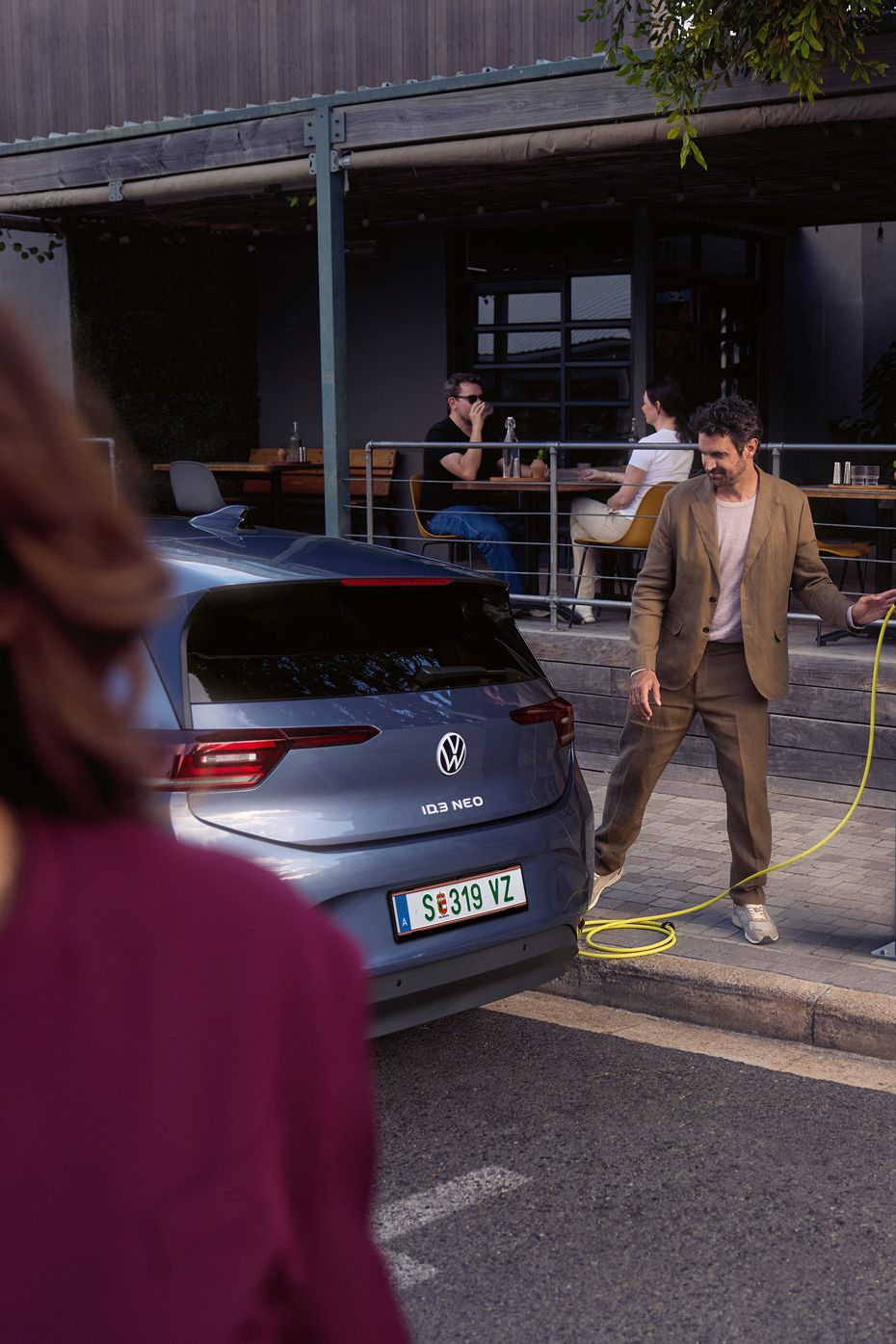 Eine Frau geht auf einer Straße. Hinter ihr läd ein Mann einen VW ID.3 Neo an einer Ladesäule vor einem Café