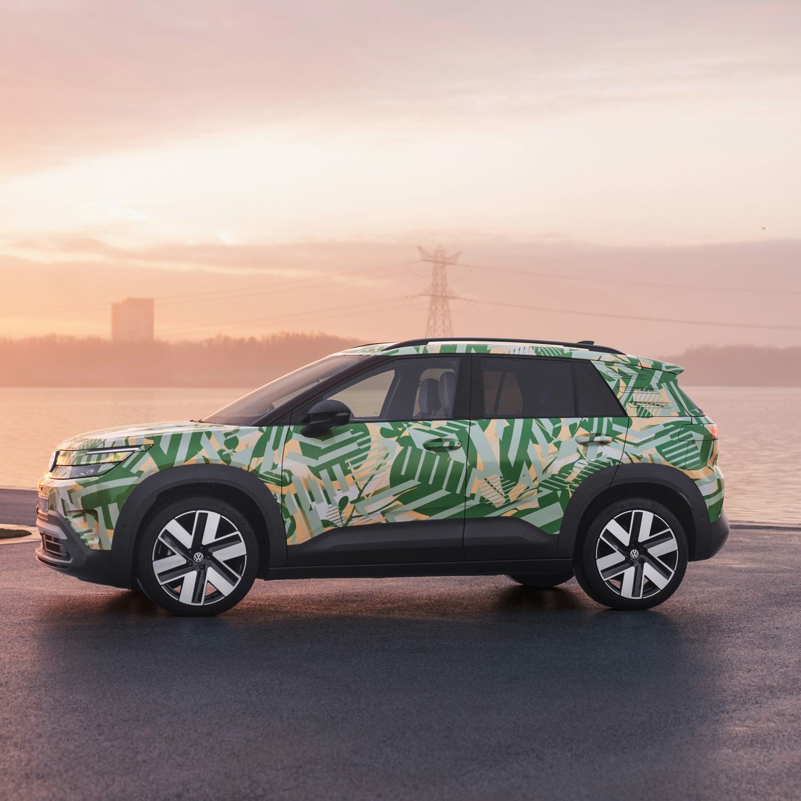 Ein SUV mit einer grünen und weißen Tarnlackierung steht auf einem asphaltierten Platz vor einem Gewässer bei Sonnenuntergang. Im Hintergrund sind Strommasten und Gebäude zu sehen. Der Himmel ist leicht bewölkt und in warmen Farben gehalten.