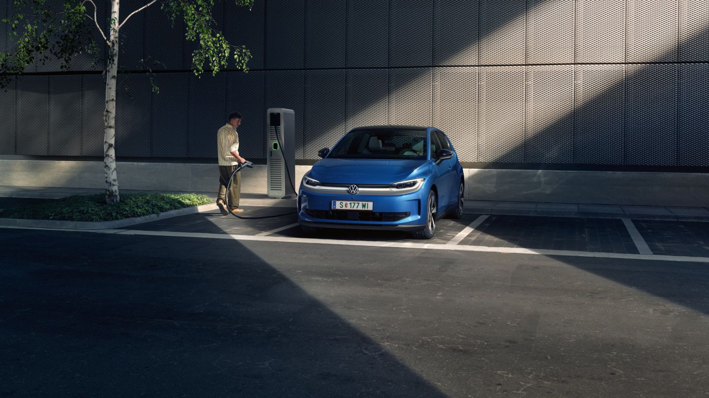 Ein blauer ID. Polo steht auf einem Parkplatz neben einer Ladesäule. Im Hintergrund sind ein Gebäude und ein Baum zu sehen.