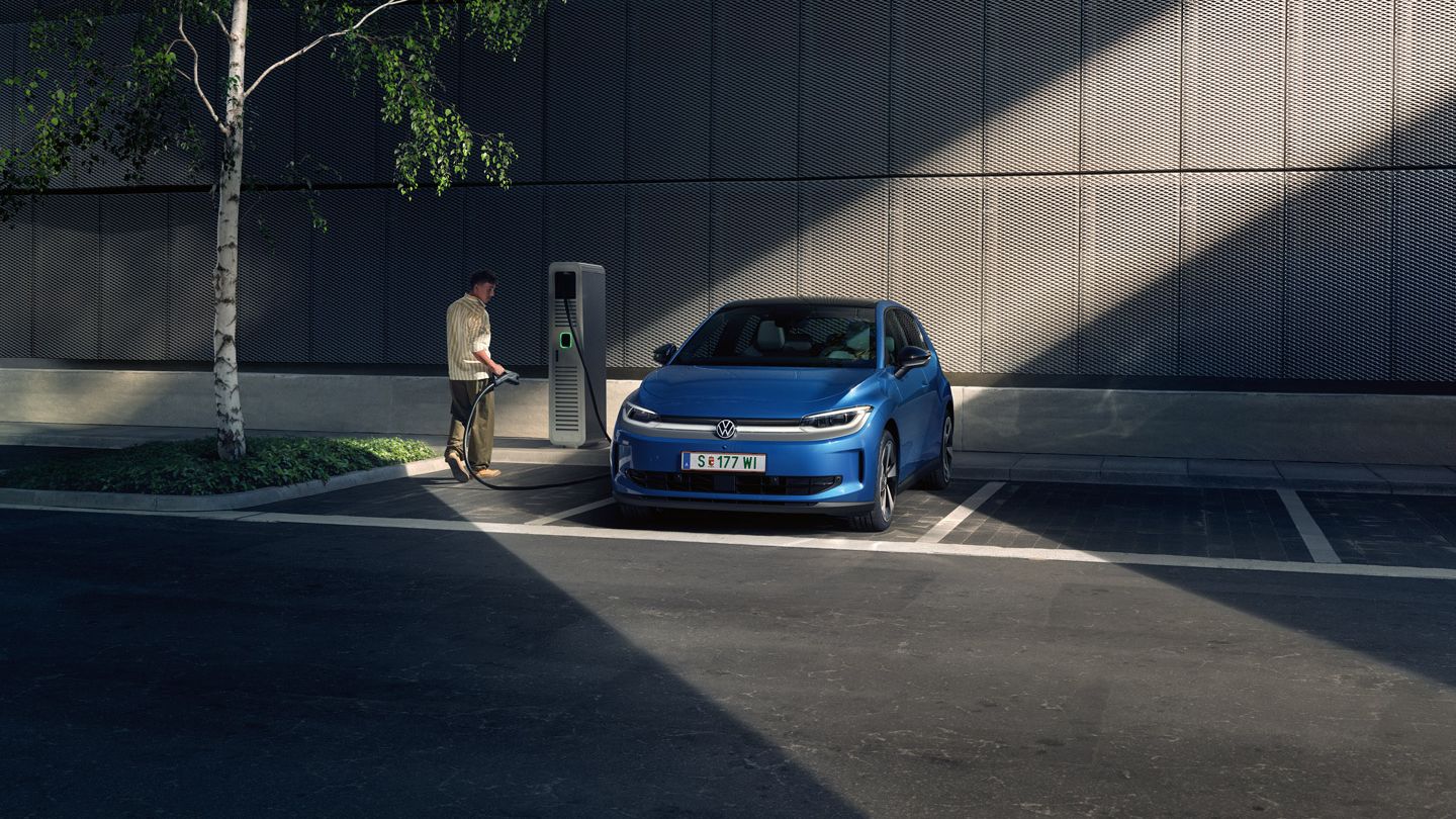 Ein blauer ID. Polo steht auf einem Parkplatz neben einer Ladesäule. Im Hintergrund sind ein Gebäude und ein Baum zu sehen.