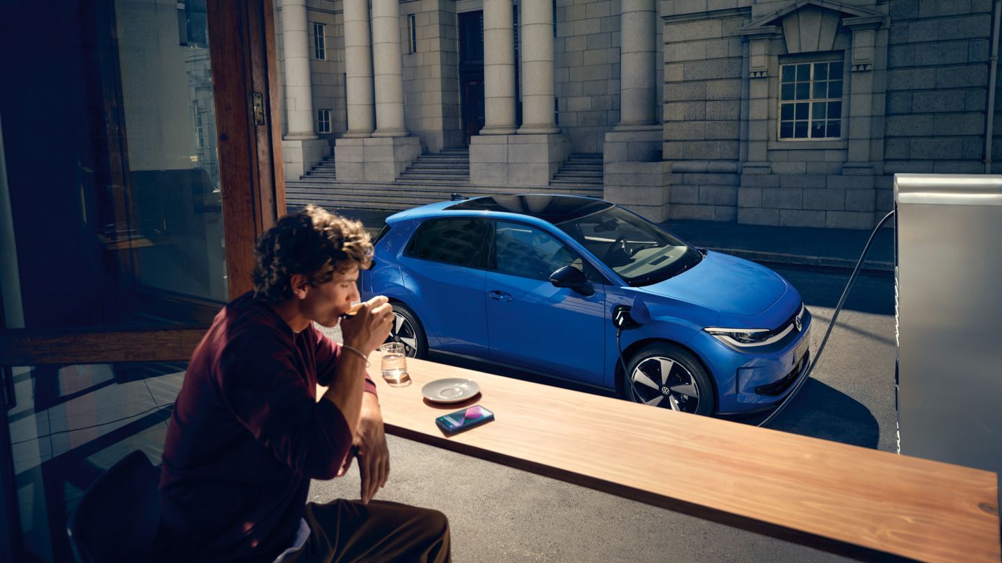 Ein Mann sitzt in einem Café, durch ein offenes Fenster ist der blaue VW ID. Polo Style zu sehen, der an einer Ladestation lädt.