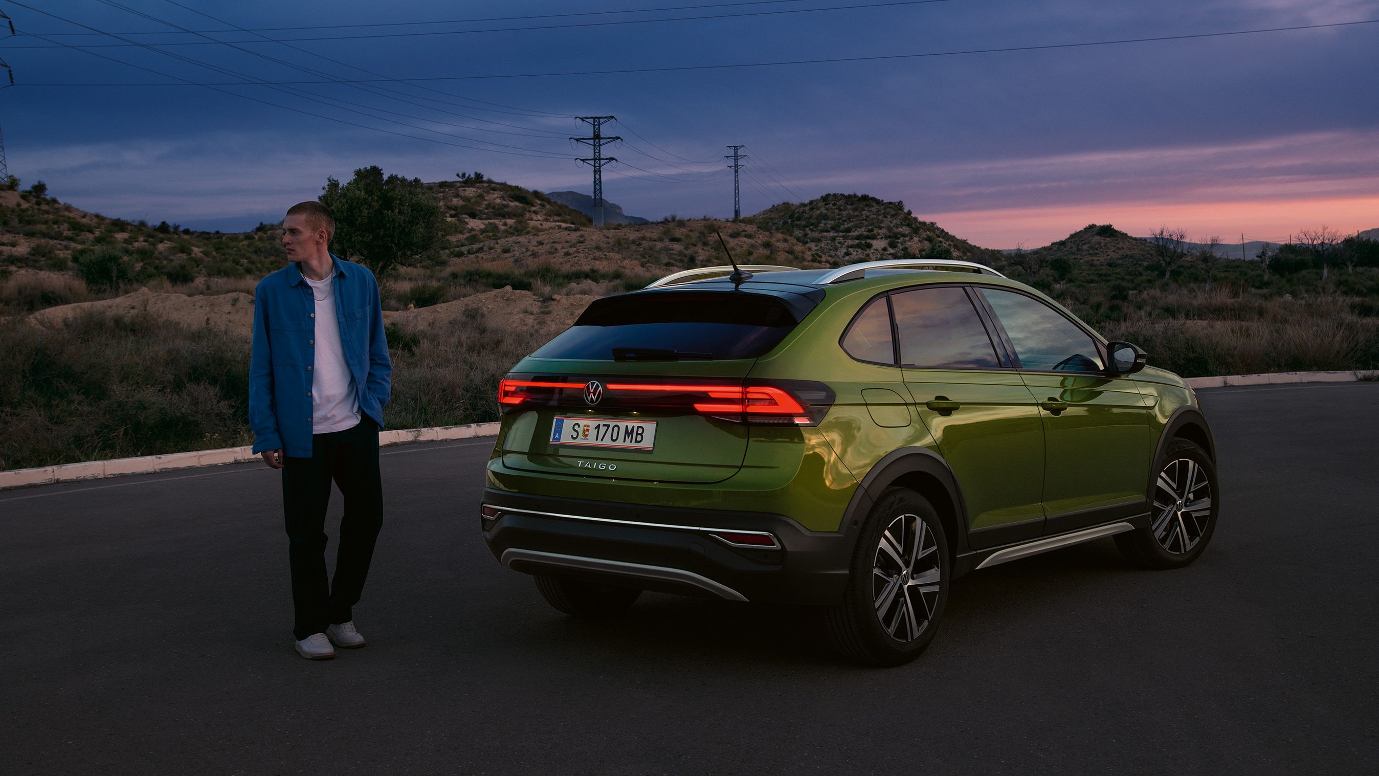 Ein Mann steht hinter einem grünen VW Taigo mit eingeschaltetem Licht auf einem Wüstenparkplatz im Sonnenuntergang.