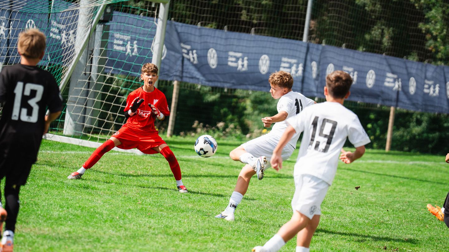 Mehrere Kinder spielen Fußball auf einem grünen Rasenplatz, ein Spieler in Weiß schießt den Ball aufs Tor. Der Torwart in roter Kleidung steht breitbeinig vor dem Tor und versucht den Schuss zu parieren. Im Hintergrund sind weitere Spieler, ein Tornetz und eine Bande mit Werbung zu sehen.