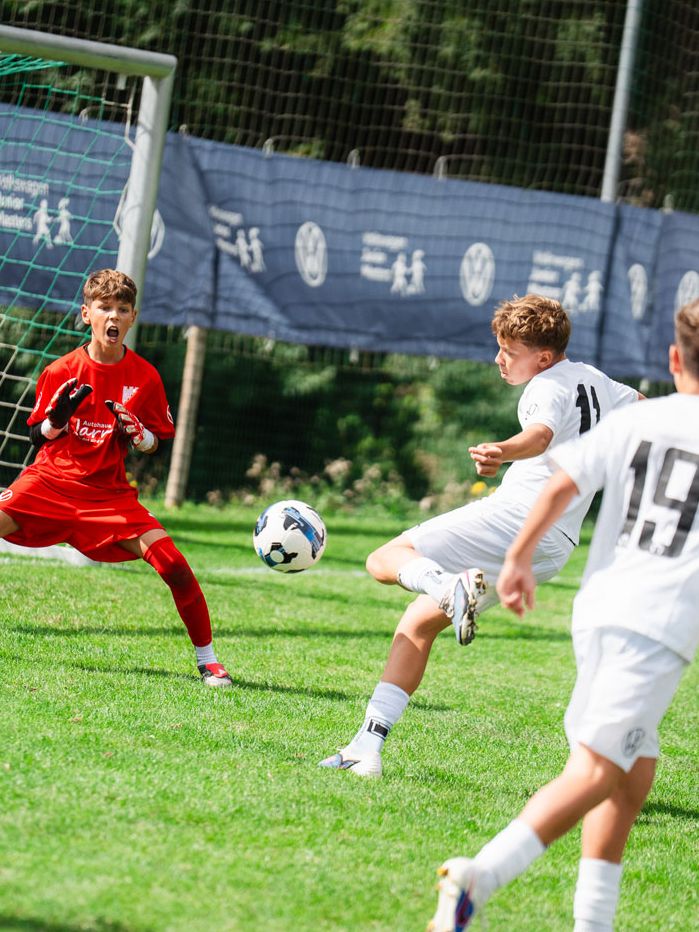 Mehrere Kinder spielen Fußball auf einem grünen Rasenplatz, ein Spieler in Weiß schießt den Ball aufs Tor. Der Torwart in roter Kleidung steht breitbeinig vor dem Tor und versucht den Schuss zu parieren. Im Hintergrund sind weitere Spieler, ein Tornetz und eine Bande mit Werbung zu sehen.