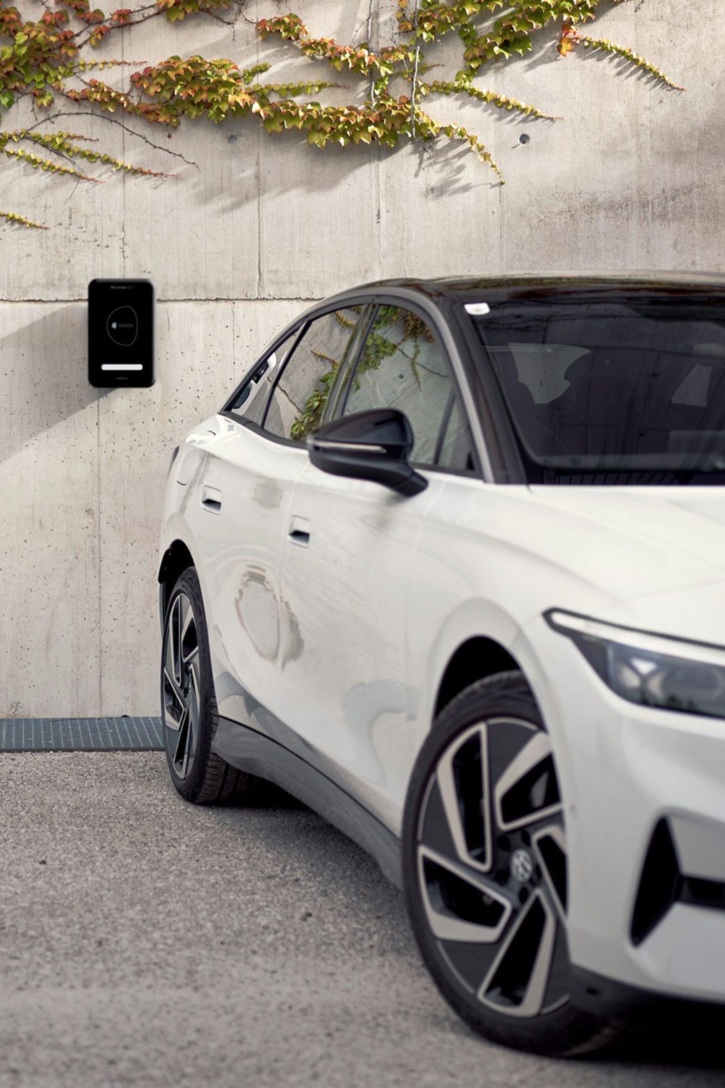 Eine Frau geht in der Nähe eines weißen ID.7, das an einer Wand mit einer Ladestation und Kletterpflanzen geparkt ist. Die Umgebung wirkt urban und modern.