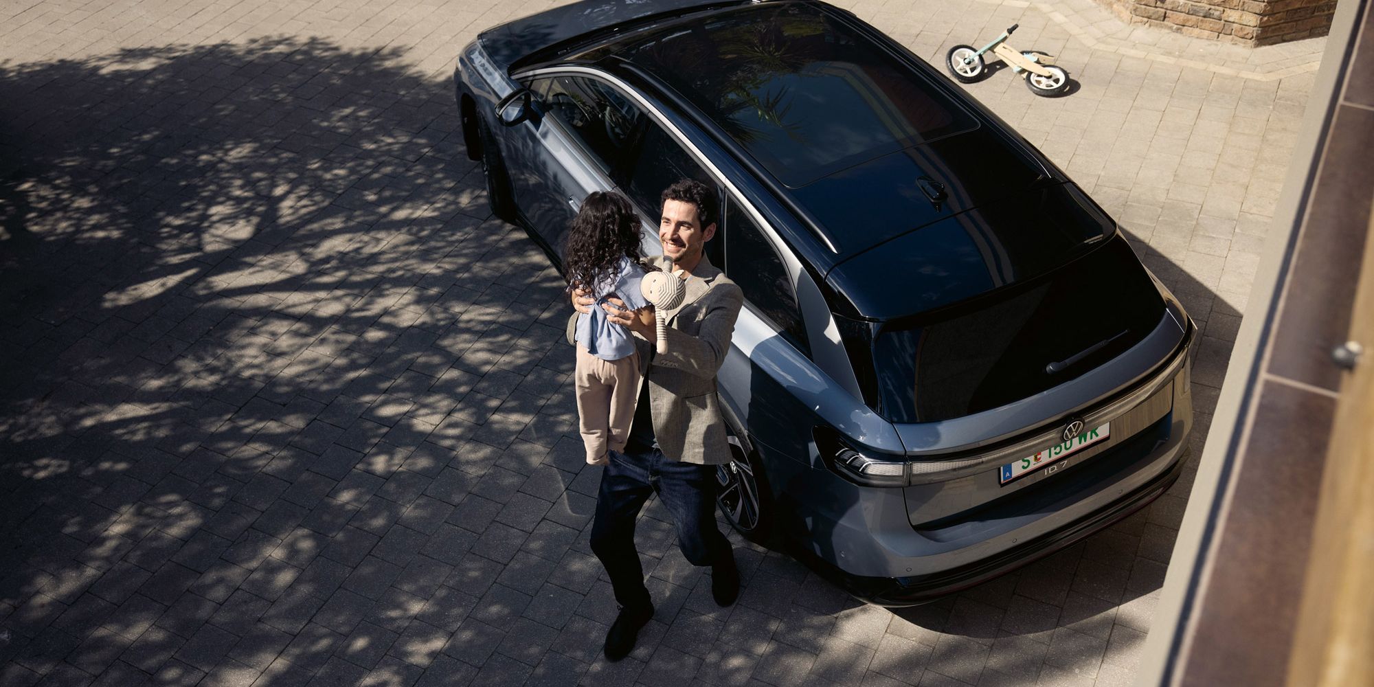 Mann hebt sein Kind hoch, was eine Puppe in der Hand halt und dahinter steht geparkt in der Einfahrt ein ID.7 Tourer und ein umgekipptes Kinderfahrrad.