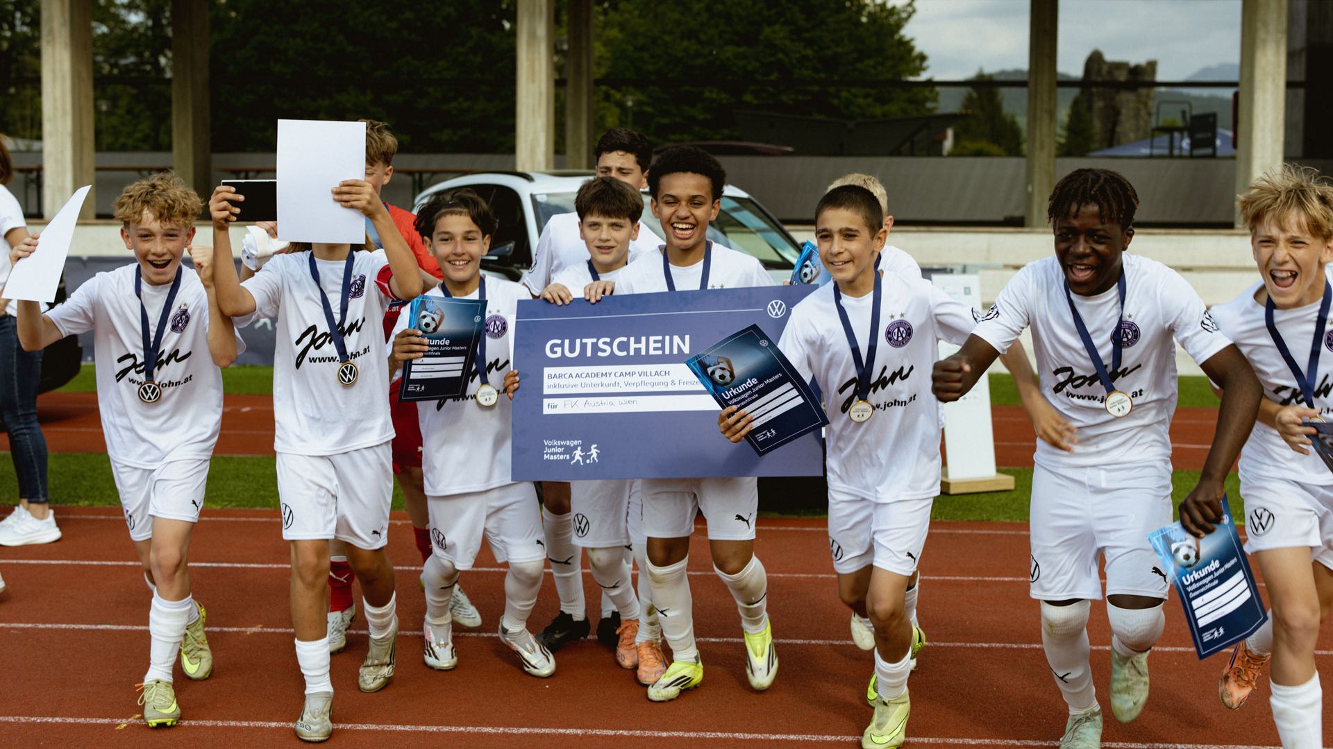 Eine Gruppe von Jungen in weißen Fußballtrikots läuft jubelnd über eine Laufbahn, viele tragen Medaillen um den Hals. Ein paar Kinder halten einen großen blauen Gutschein sowie Urkunden in den Händen. Im Hintergrund sind ein Auto, eine Tribüne und Gebäude zu sehen.