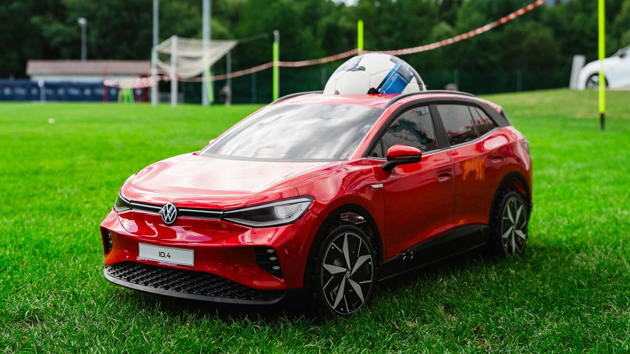 Ein rotes Modellauto im Stil eines modernen VW-SUV steht auf einer grünen Wiese. Im Hintergrund sind ein Sportplatz mit Flutlichtmast, Bäume und Zelte zu sehen. Der Himmel ist leicht bewölkt, was der Szene ein natürliches Licht verleiht.