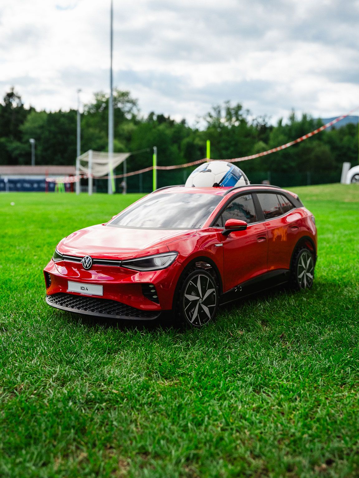 Ein rotes Modellauto im Stil eines modernen VW-SUV steht auf einer grünen Wiese. Im Hintergrund sind ein Sportplatz mit Flutlichtmast, Bäume und Zelte zu sehen. Der Himmel ist leicht bewölkt, was der Szene ein natürliches Licht verleiht.