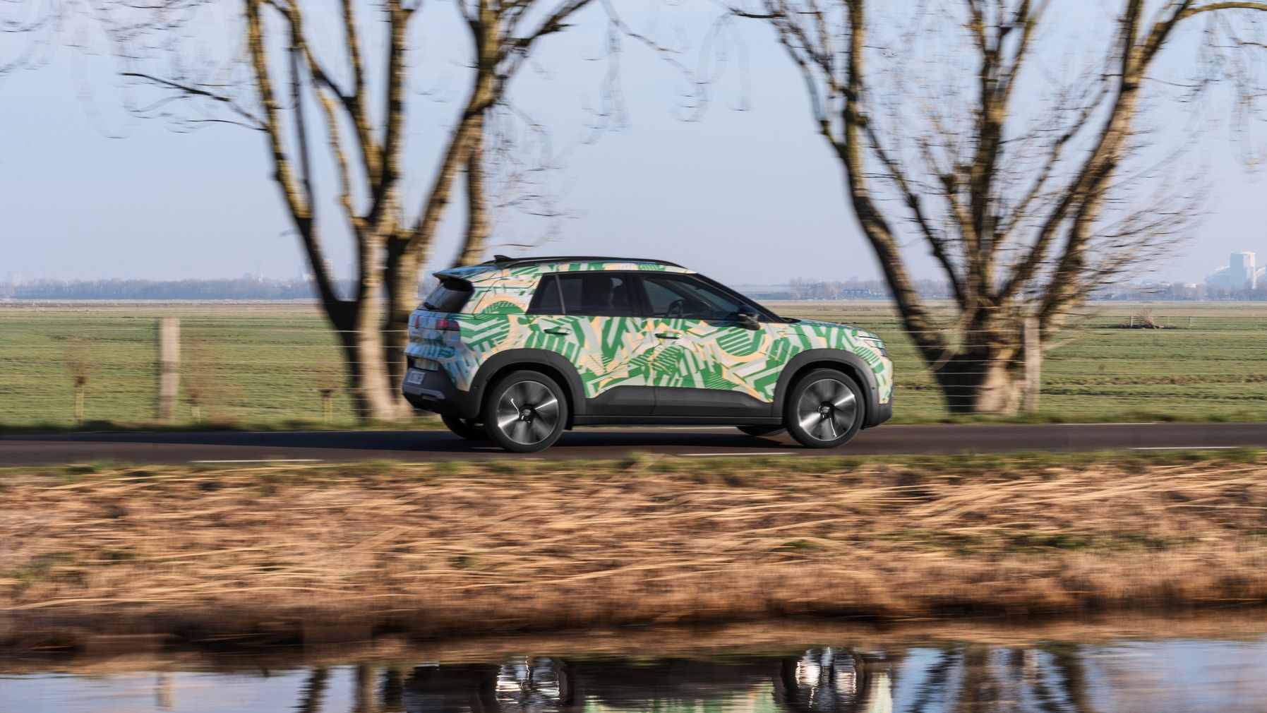 Ein SUV fährt auf einer ländlichen Straße vorbei an kahlen Bäumen und einem Wassergraben. Das Auto ist mit einem bunten, tropischen Muster bemalt. Im Hintergrund ist eine grüne Wiese und wolkenloser Himmel zu sehen.