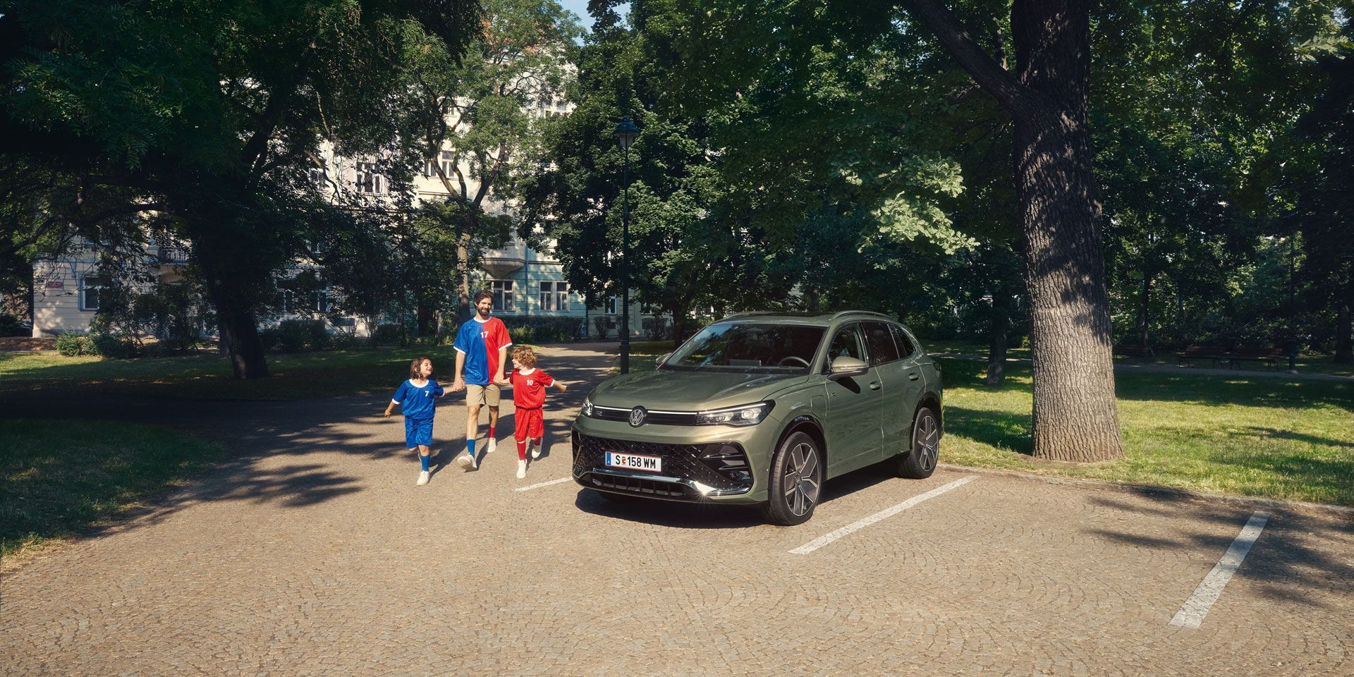 Ein Mann geht mit zwei Kindern von einem VW Tiguan R-Line Plug-In- Hybrid weg, der im Park steht.