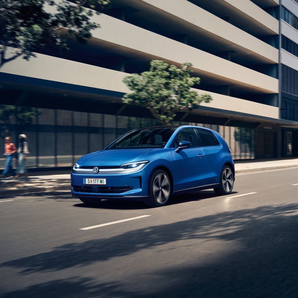 Ein blauer VW ID. Polo Style fährt durch eine Stadt mit grünen Bäumen am Straßenrand.