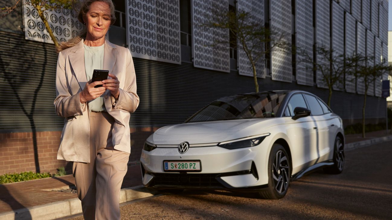 Eine Frau im Anzug schaut auf ihr Smartphone, im Hintergrund parkt ein weißer VW ID.7 am Straßenrand.