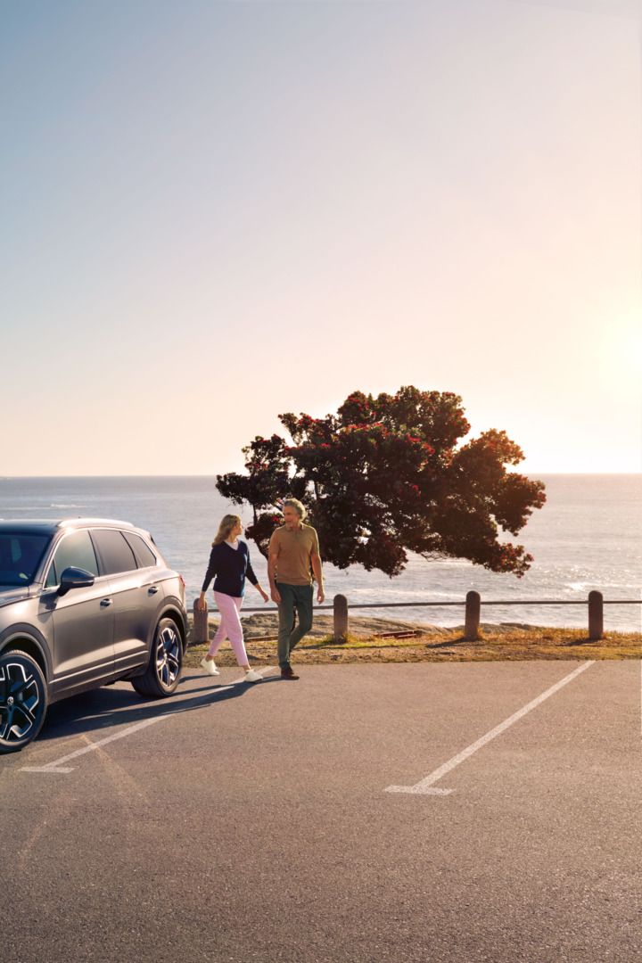 Der neue Volkswagen Touareg auf einem Parkplatz an der Küste, an ihm gehen eine Frau und ein Mann vorbei