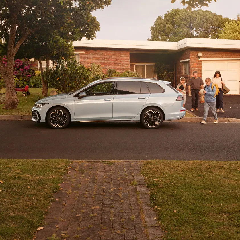 Ein weißer VW Golf Variant parkt vor einem Bungalow in einer Wohnstraße mit Bäumen. 