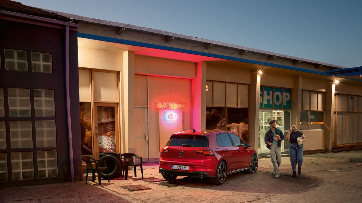 Roter VW Golf GTI steht abends vor einem Supermarkt, zwei junge Leute gehen mit Einkäufen daran vorbei.
