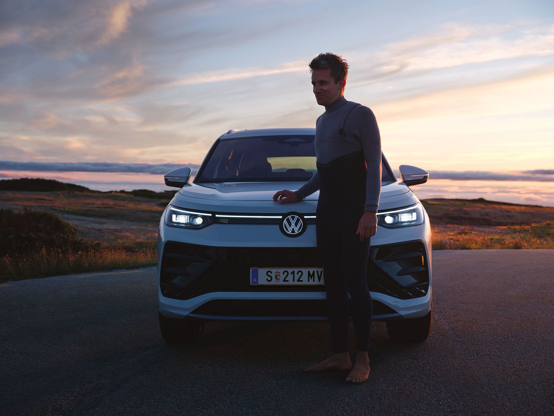 Mann im Neoprenanzug steht im Sonnenuntergang an der Motorhaube eines VW Tayron mit aktiver Lichtsignatur.