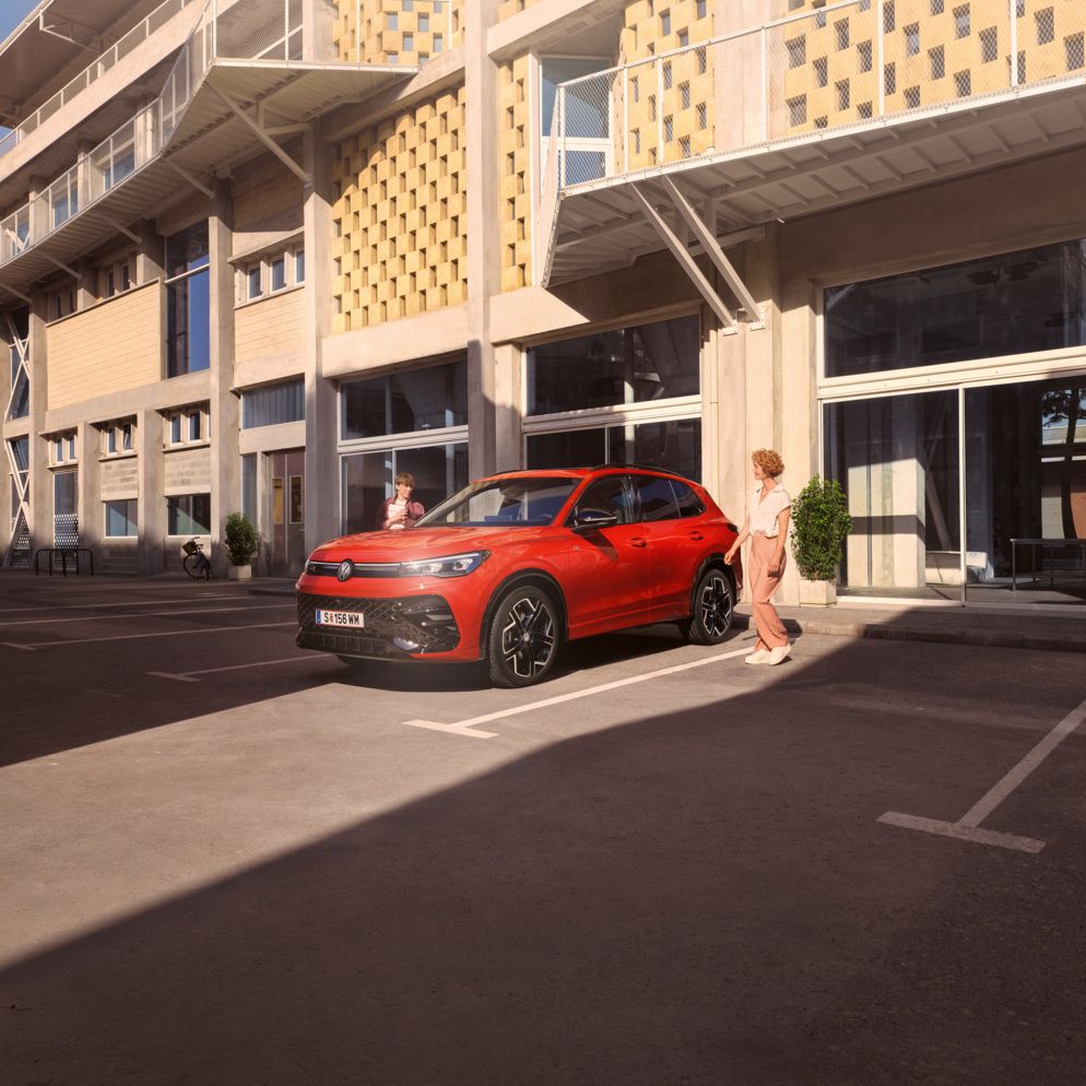 Roter VW Tiguan R-Line in Seitenansicht steht vor einem Gebäude, eine junge Frau geht auf der Fahrerseite vorbei.