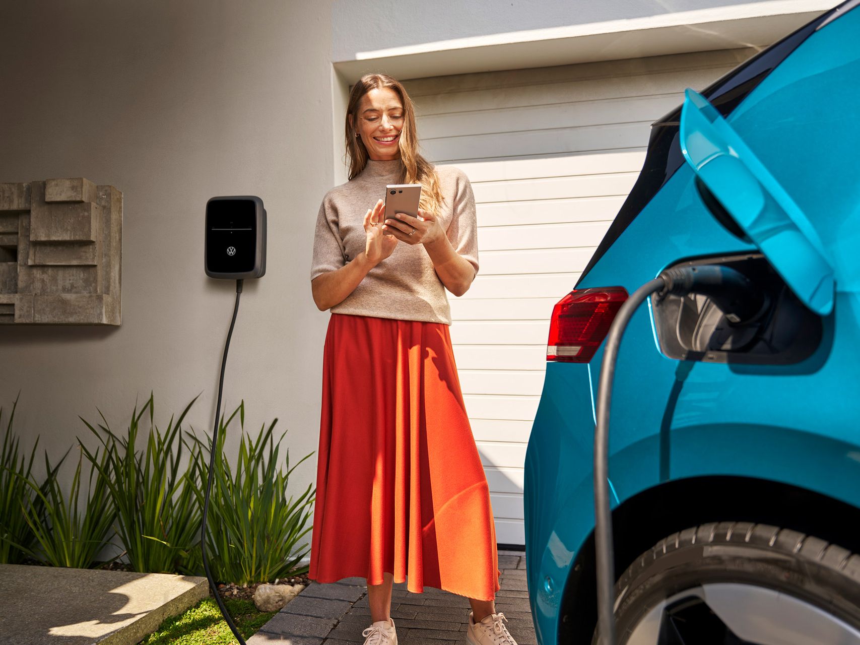 Eine Frau steht vor einer Garage und bedient lächelnd ihr Smartphone, während neben ihr ein blaues Elektroauto an einer Wallbox geladen wird. Das Ladekabel ist deutlich am Auto angeschlossen, und im Hintergrund sind Pflanzen und die Hausfassade zu sehen.