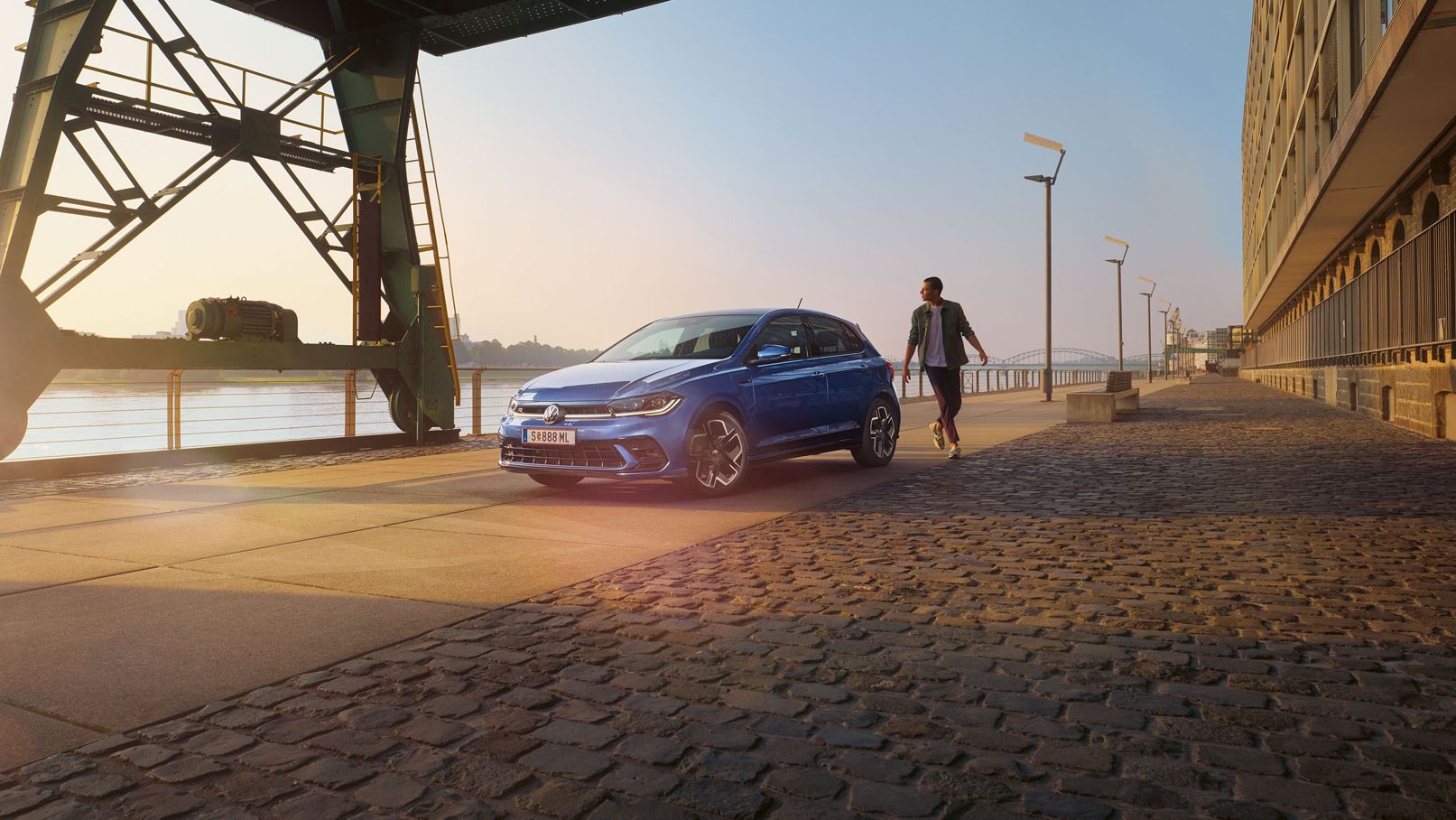  Ein blauer VW Polo R-Line steht an der Promenade neben einem Fluss, ein Mann läuft um das Auto herum.