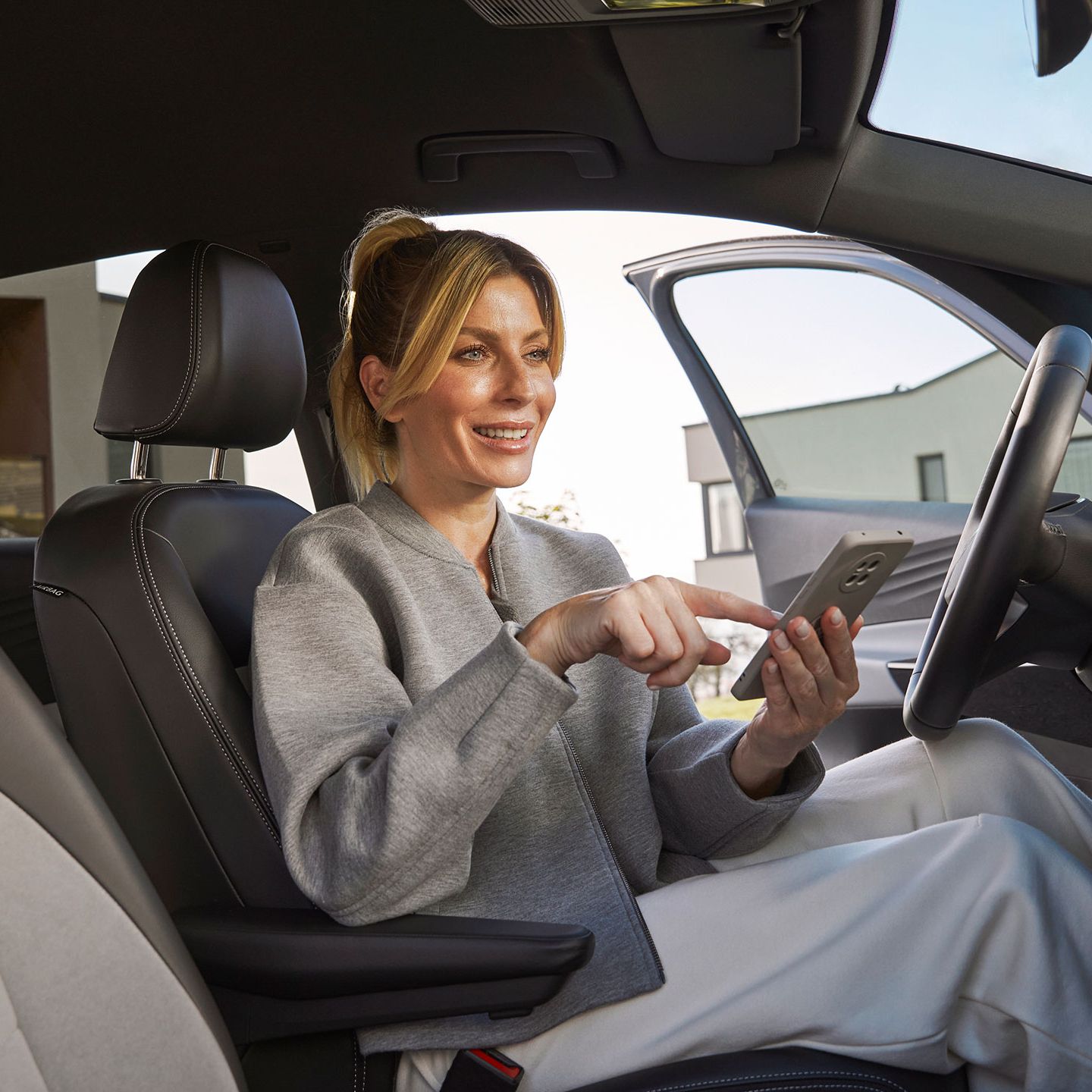 Eine Frau sitzt in einem Volkswagen auf dem Fahrersitz und bedient ein Smartphone. Der Innenraum des Autos wirkt hochwertig. 