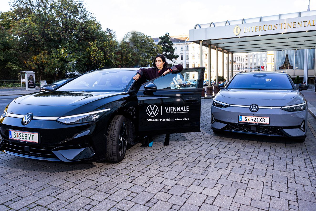 Eine Person steht vor dem Eingang des InterContinental Hotels neben einem schwarzen Volkswagen-Elektroauto und hält die geöffnete Fahrertür. Rechts daneben steht ein zweites, graues Elektroauto desselben Modells. Auf der Tür des schwarzen Wagens ist ein Logo der Viennale mit dem Hinweis „Offizieller Mobilitätspartner 2024“ zu sehen.