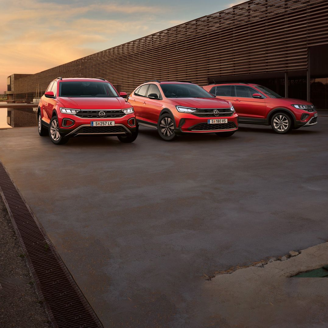 T-Roc, Taigo, T-Cross sind nebeneinander auf einem Parkplatz bei Sonnenuntergang aufgereiht. Im Hintergrund befindet sich ein modernes Gebäude. Der Himmel zeigt eine sanfte Abenddämmerung.