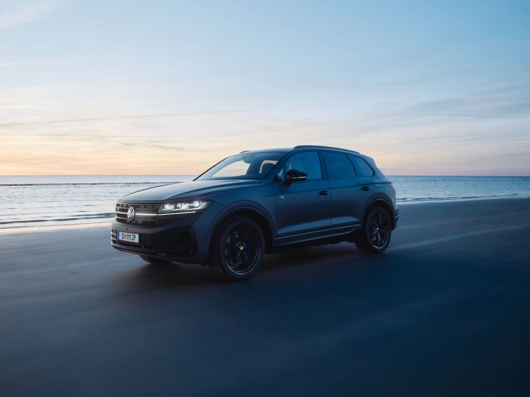 Ein dunkler Touareg R fährt bei Sonnenuntergang auf einem Strand neben dem Meer. Der Himmel ist überwiegend blau, mit einigen orangefarbenen und gelben Tönen der untergehenden Sonne. Das Meer ist ruhig im Hintergrund.