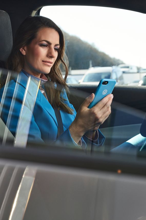 Eine in blau gekleidete Frau sitzt in einem geparkten Volkswagen mit einem Smartphone in ihrer Hand.