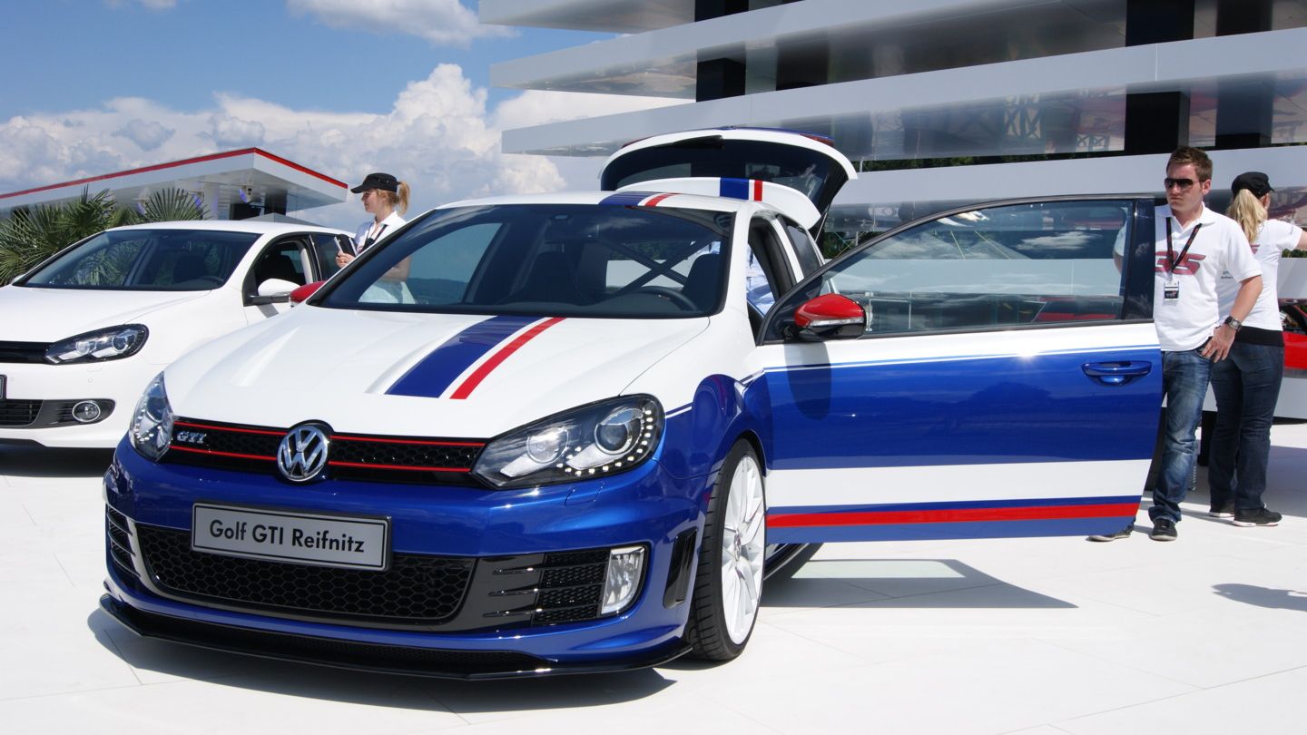 Ein blauer Volkswagen Golf GTI mit weißen und roten Streifen steht auf einem Messegelände. Das Auto hat geöffnete Türen, und im Hintergrund sind weitere Fahrzeuge und Gebäude zu sehen. Eine Person steht neben dem Auto.